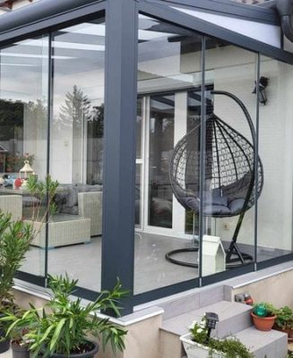 Seră de grădină închisă în sticlă, cu scaun suspendat tip cuib și plante decorative, protejată de rulou exterior